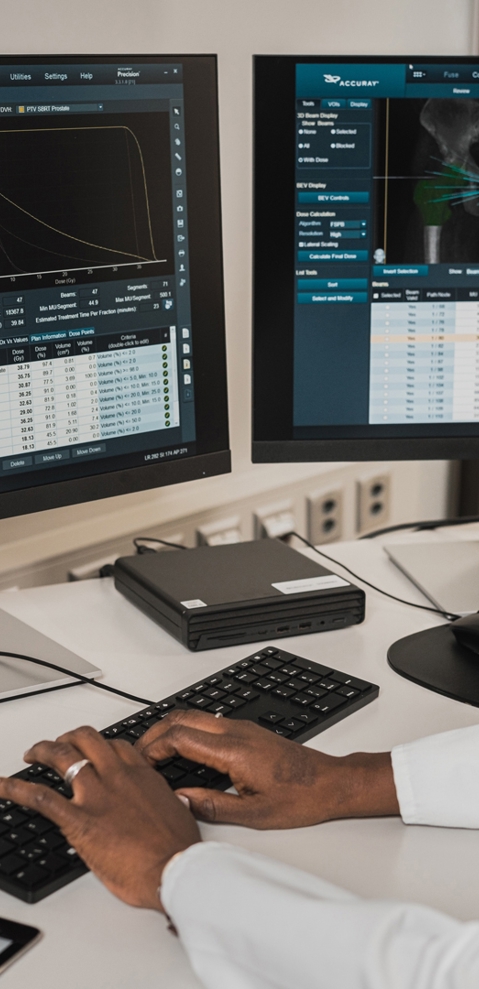 A radiologist sitting at a desk, looking at two computer screens showing scan software